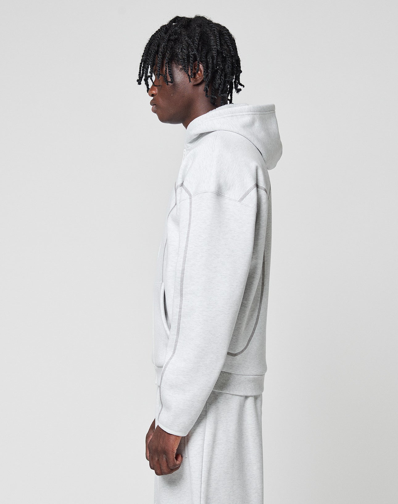 A person with short dreadlocks wears the LIVE FAST DIE YOUNG LFDY Active Fleece Hooded Zip in an oversize fit, light gray with subtle logo print, paired with matching sweatpants, standing in profile against a white background.