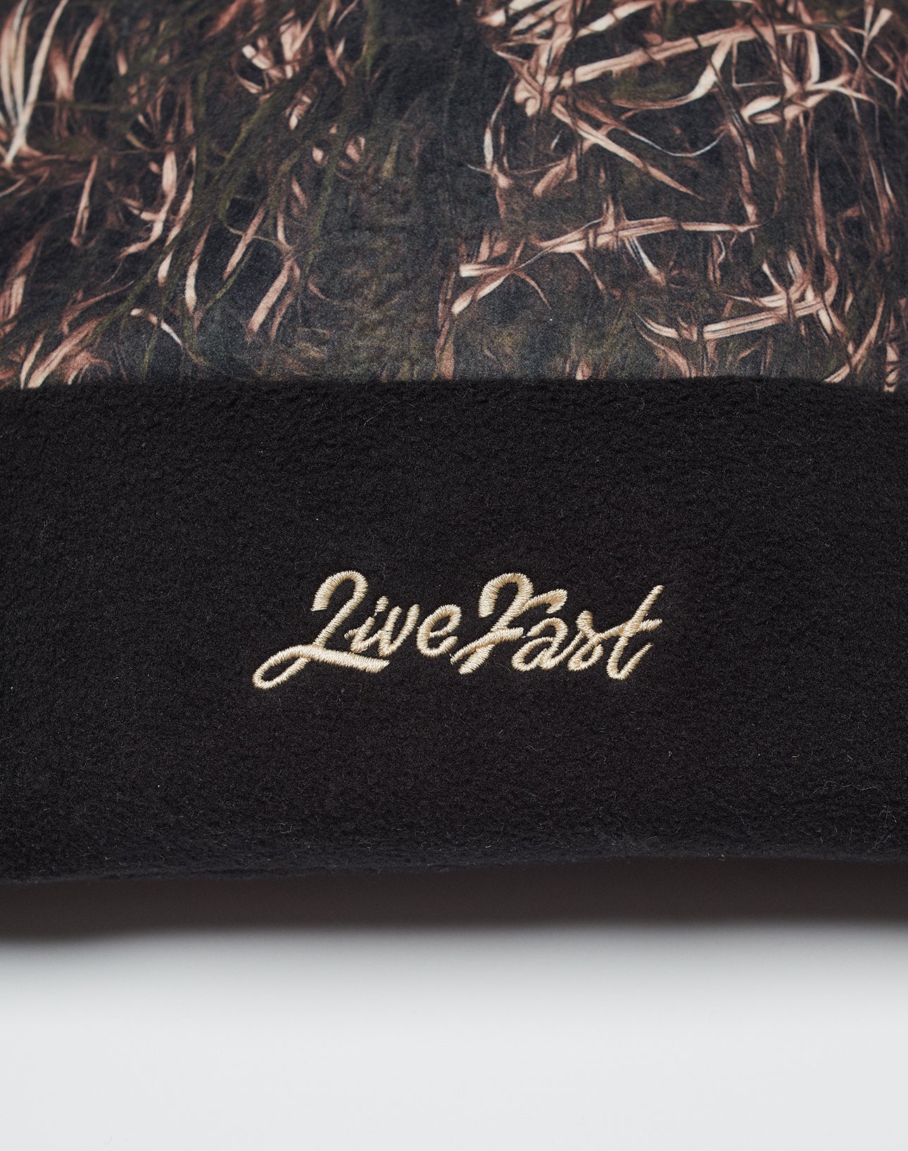 A close-up of the LIVE FAST DIE YOUNG Live Fast Gymbag reveals brown and green camo fabric on top, black fleece on the bottom, and LiveFast embroidered in beige script on the black polyester section.