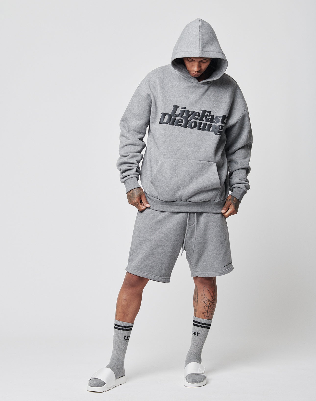Wearing an oversize fit Basic Hooded by LIVE FAST DIE YOUNG, gray shorts and socks, and white slide sandals, a person stands against a plain white background with hands in pockets, head slightly down, and hood up.
