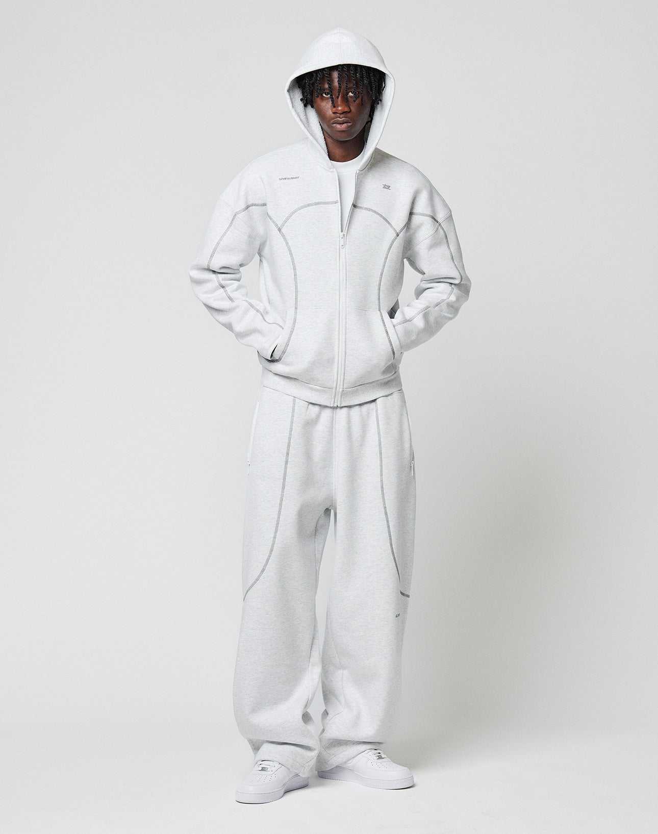 A person stands against a plain background wearing LIVE FAST DIE YOUNGs LFDY Active Fleece Jogger in light gray with a matching hooded zip-up sweatshirt, Reißverschlusstaschen, white sneakers, hands in pockets, and the hood up.