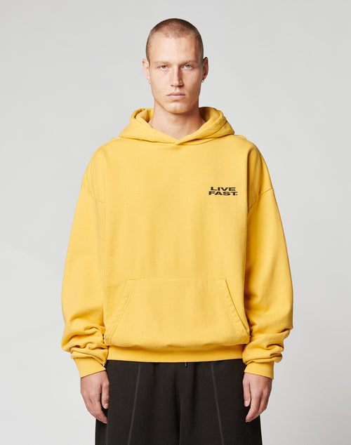 A person with short hair wears a loose-fit yellow Print Over Hooded hoodie by LIVE FAST DIE YOUNG, featuring LIVE FAST in black on the left chest, standing against a plain light gray background.