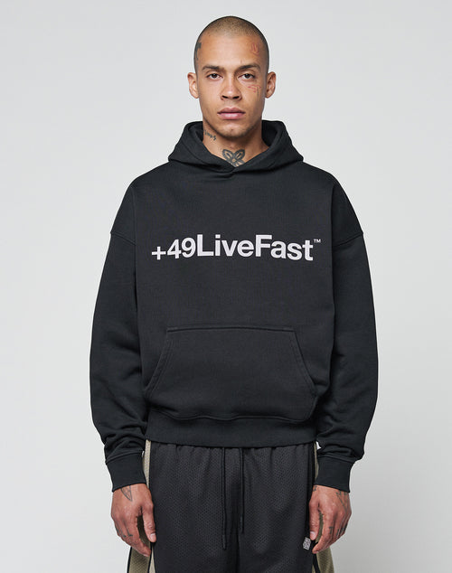 A person with a shaved head and neck tattoos wears the LIVE FAST DIE YOUNG Basic +49 Hooded in oversize fit black, featuring a bold white +49LiveFast front print and crafted from a soft cotton-polyester blend, standing against a plain light background.