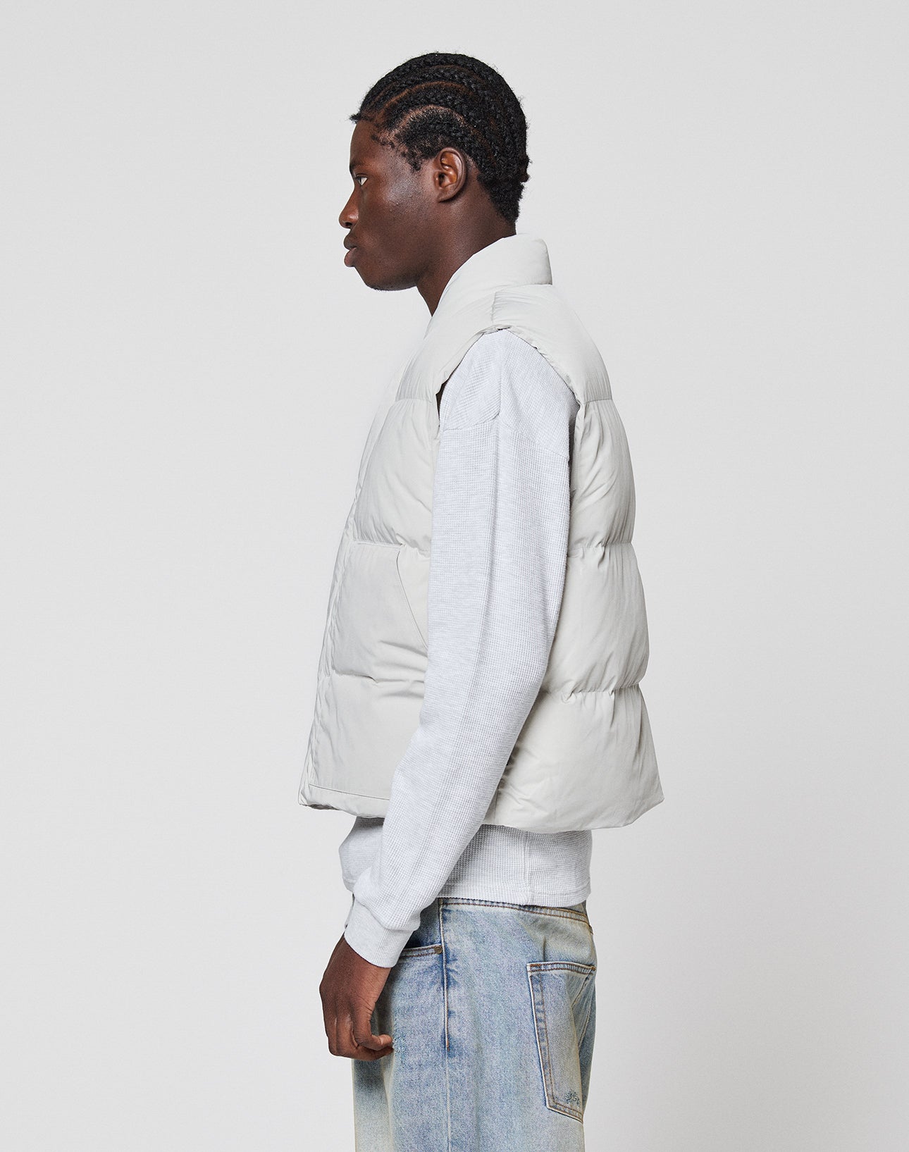 A person stands in profile against a plain white background, wearing the LIVE FAST DIE YOUNG Basic Cropped Vest in light grey polyester over a grey sweatshirt and light blue jeans.