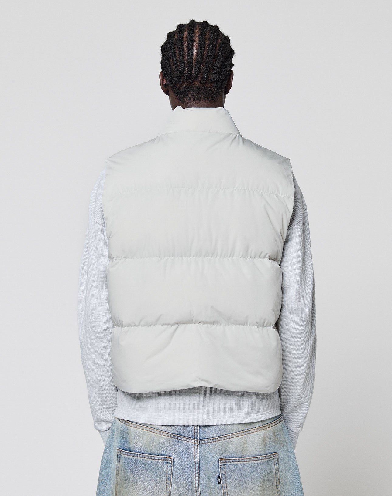 A person with braided hair, seen from behind, wears the LIVE FAST DIE YOUNG Basic Cropped Vest in light gray polyester over a gray sweatshirt and light blue jeans against a plain light background.