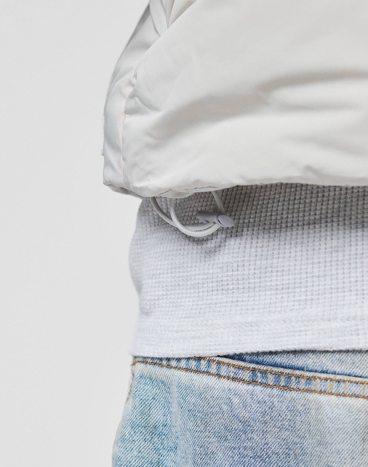 Close-up of a person wearing the LIVE FAST DIE YOUNG Basic Cropped Vest, light blue denim jeans, and a regular fit white puffer jacket with a drawstring hem. The background is plain and light-colored.