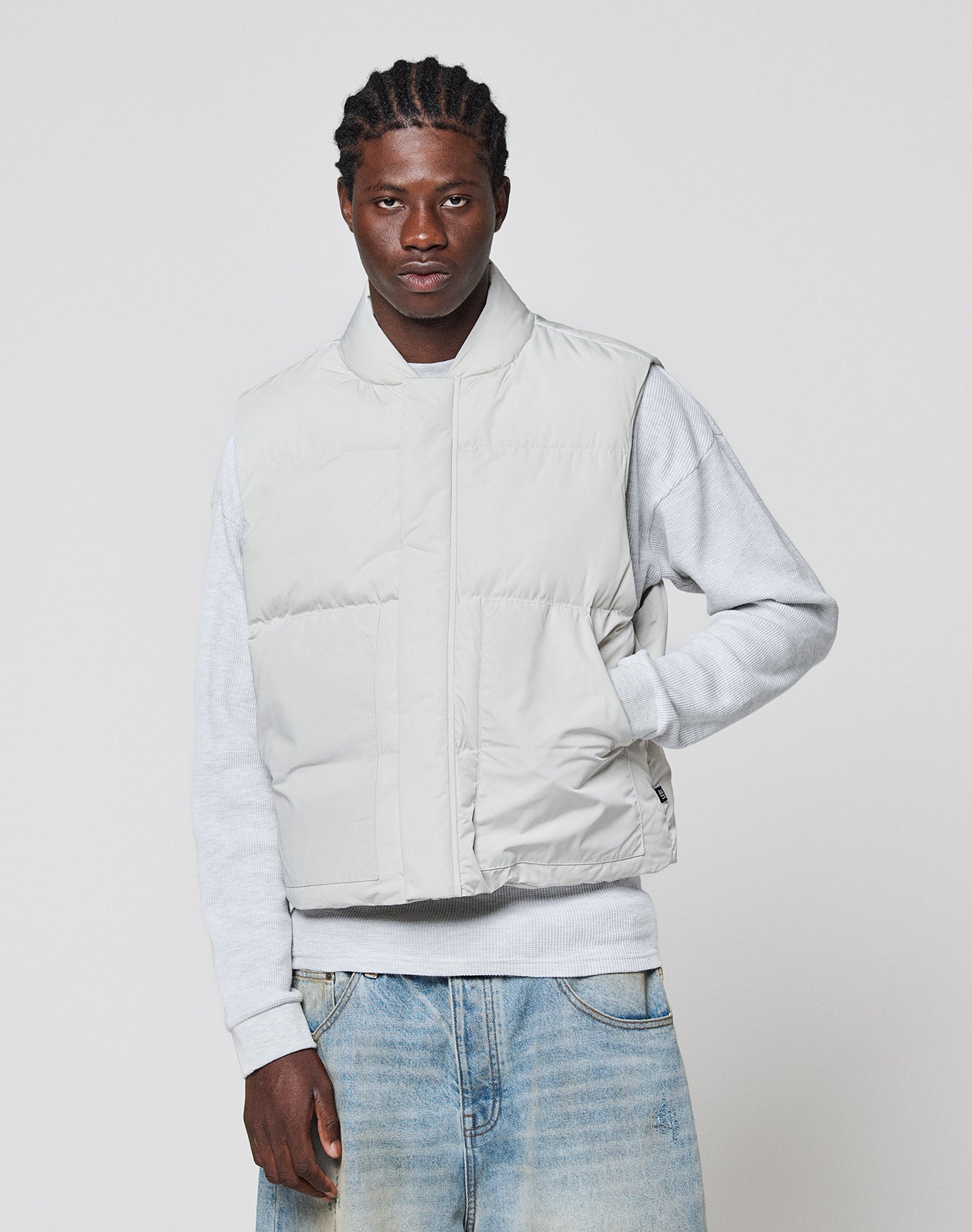 A man with braided hair wears the LIVE FAST DIE YOUNG Basic Cropped Vest in light gray over a gray sweatshirt and faded blue jeans, standing against a plain white background.