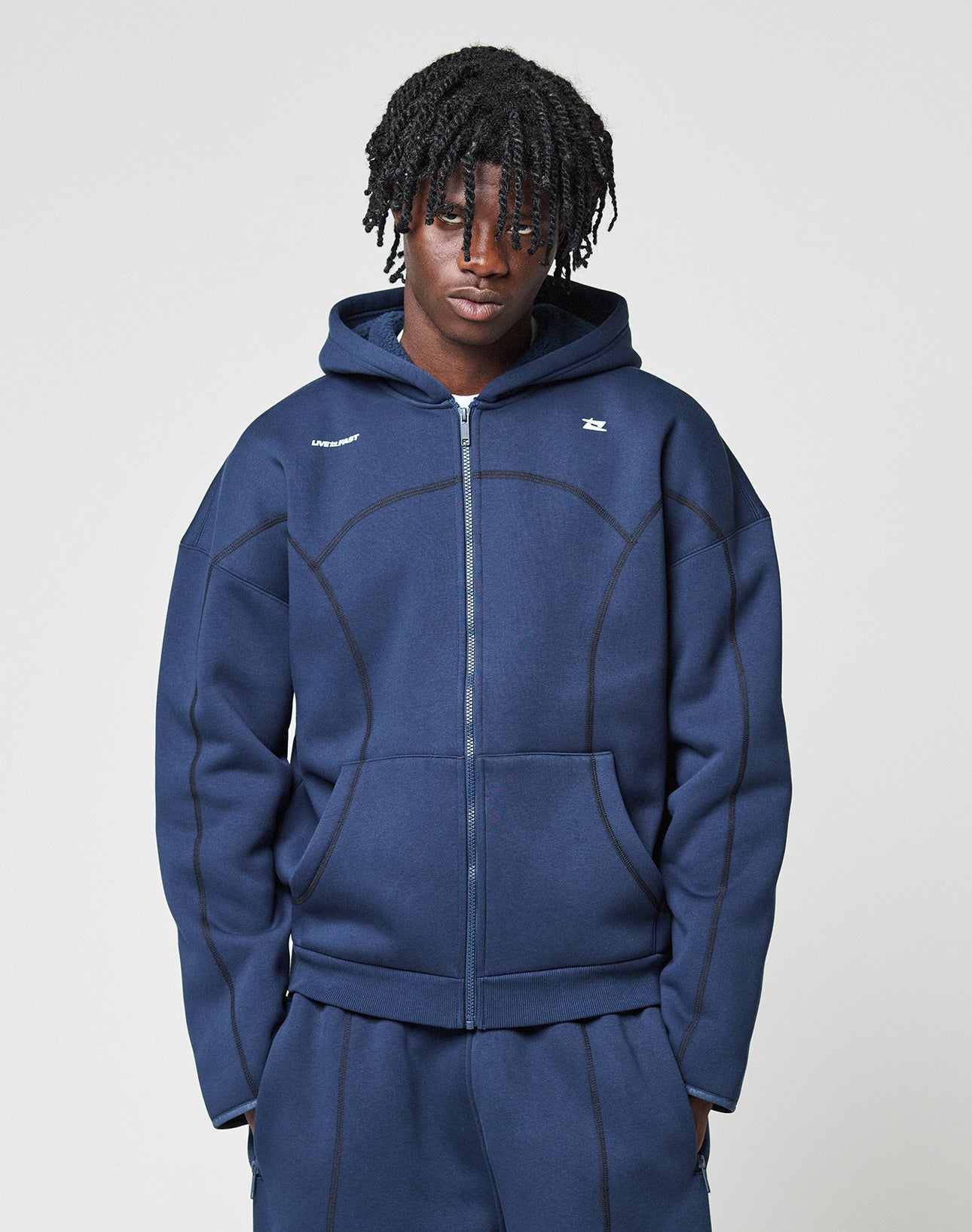 A person with medium-length, textured hair wears the LFDY Active Fleece Hooded Zip by LIVE FAST DIE YOUNG—an oversized navy zip-up hoodie with curved seam details and small white logo prints on the chest—paired with matching track pants.
