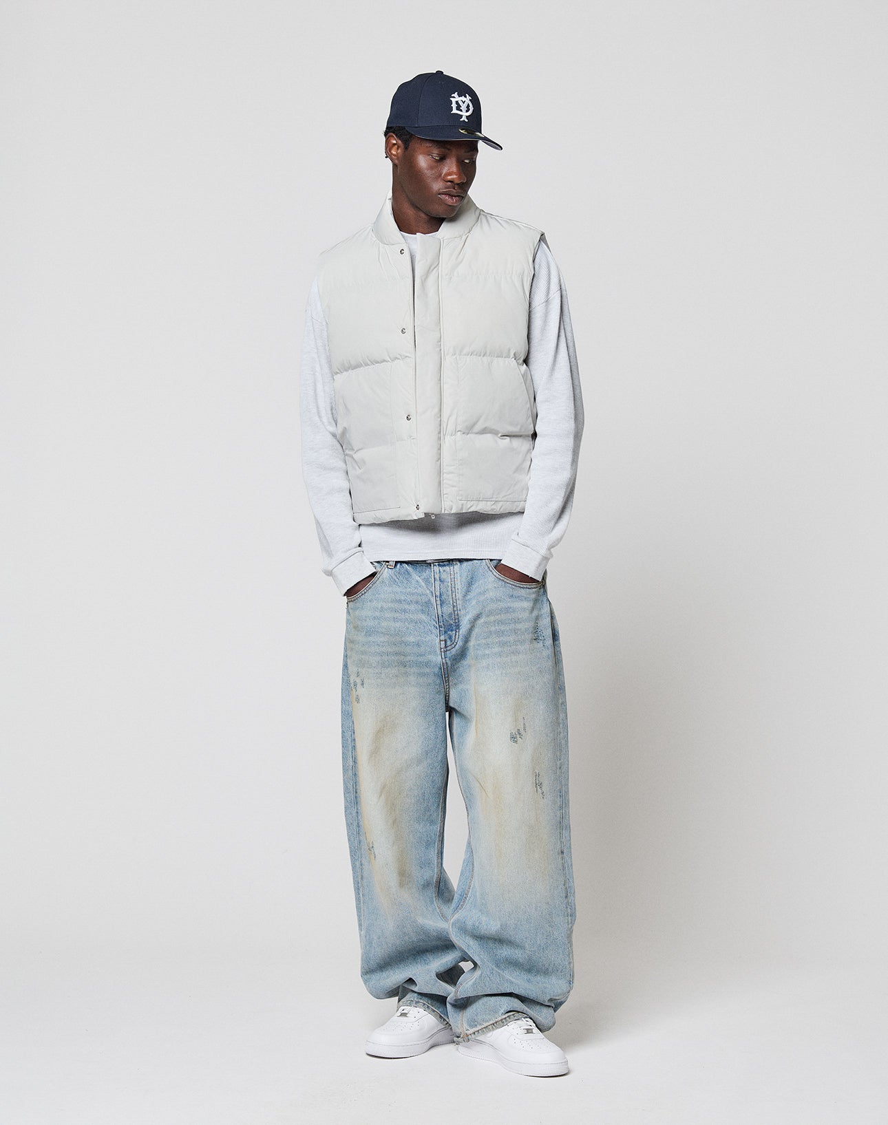 Someone wearing a LIVE FAST DIE YOUNG Basic Cropped Vest in light gray over a gray long-sleeve shirt, navy baseball cap, baggy light blue regular-fit jeans, and white sneakers stands against a plain white background.