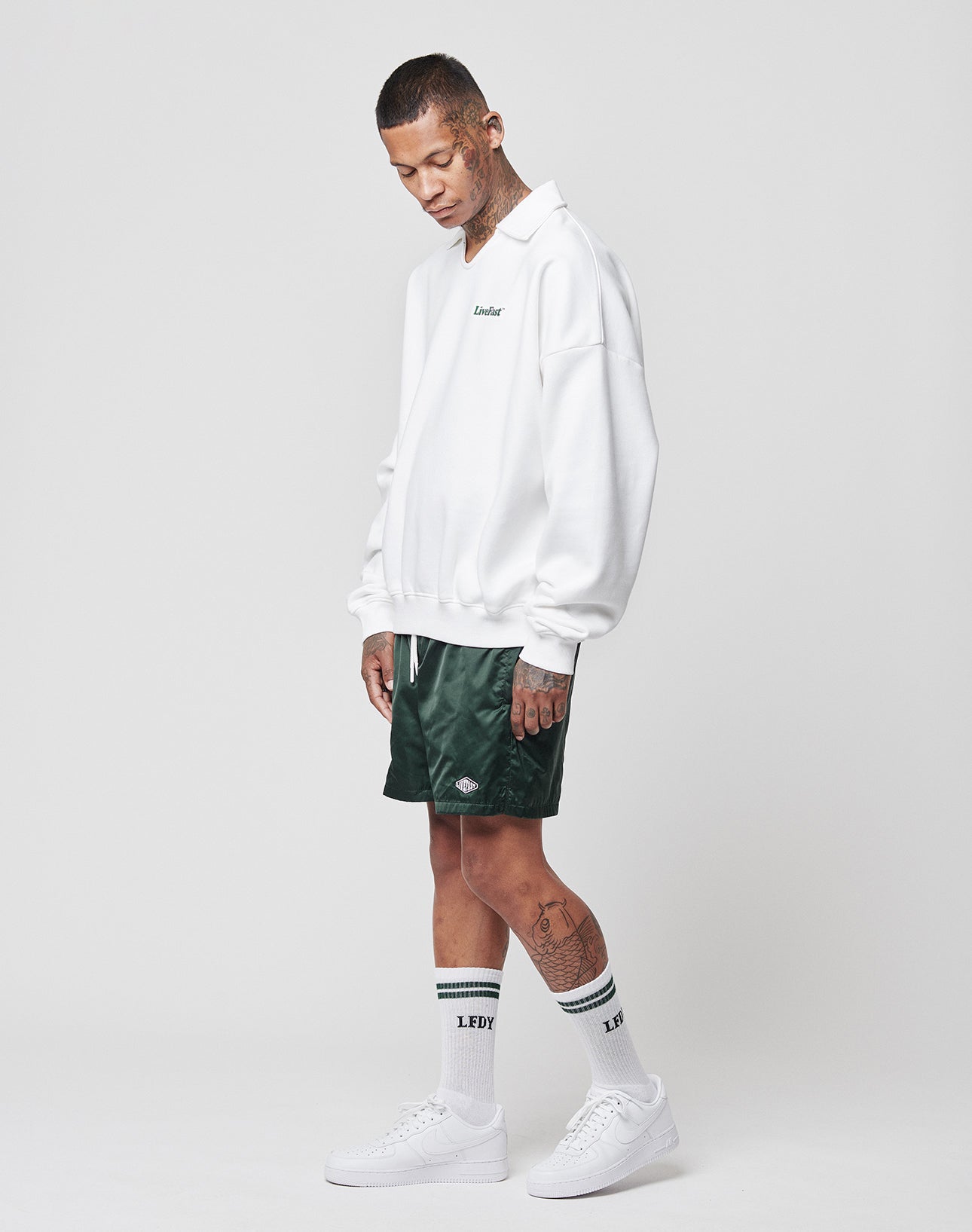 A tattooed person wears the LIVE FAST DIE YOUNG Basic Collar Sweater in an oversize fit, green athletic shorts, LFDY white crew socks, and white sneakers. They stand against a plain white background while looking downward.