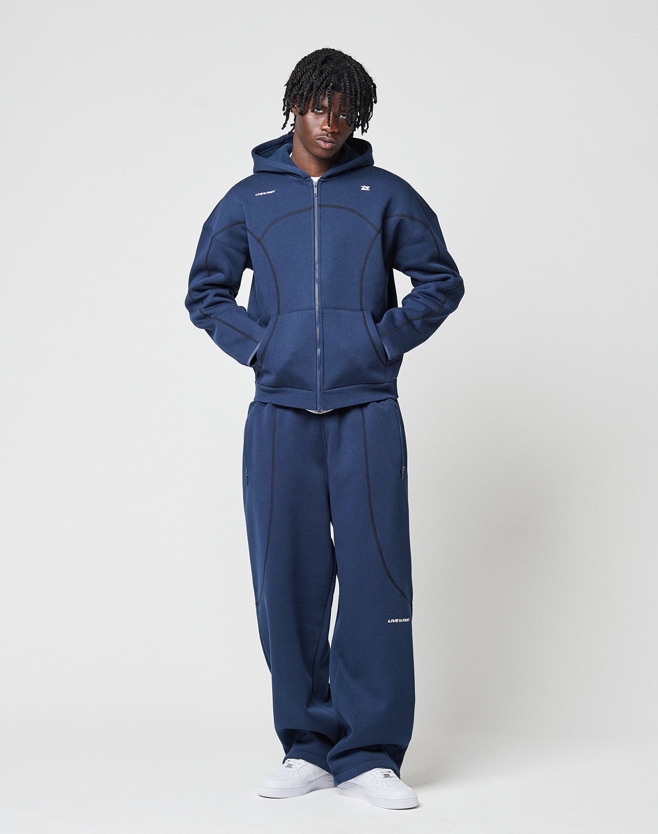A person stands against a plain white background wearing the LFDY Active Fleece Hooded Zip by LIVE FAST DIE YOUNG in navy blue with a subtle logo, paired with matching sweatpants and white sneakers. Their hands are in their pockets and they face the camera.