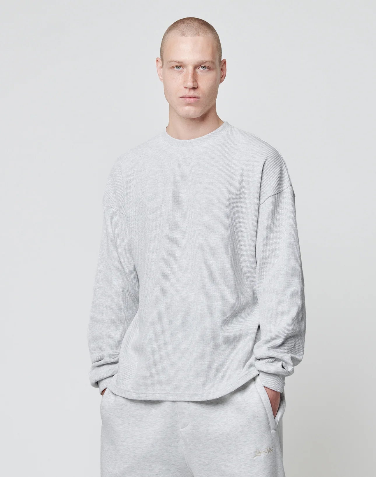 A man with short hair stands against a plain light background, wearing the LIVE FAST DIE YOUNG Blank Waffle Longsleeve in an oversize fit and matching 100% Baumwolle sweatpants, hands in pockets.