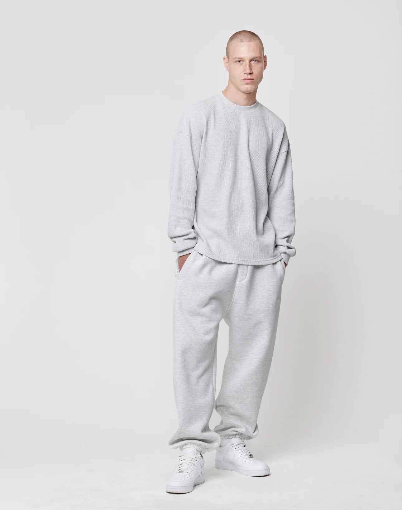 A person with a buzz cut stands against a plain white background, wearing the LIVE FAST DIE YOUNG Blank Waffle Longsleeve in an oversize fit and light gray color, hands in pockets with matching sweatpants and white sneakers.