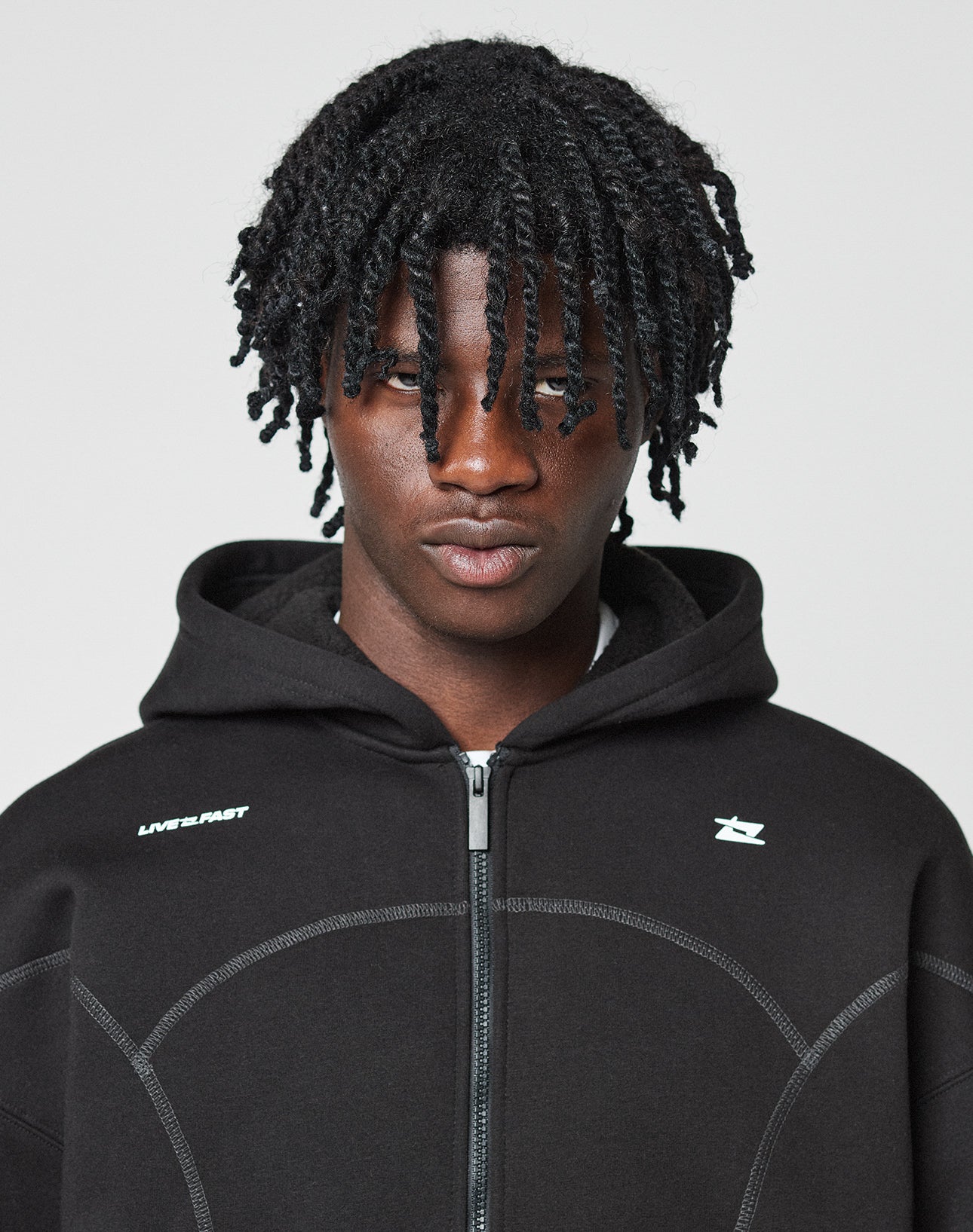 A person with medium-dark skin and tightly coiled hair looks at the camera, wearing the LIVE FAST DIE YOUNG LFDY Active Fleece Hooded Zipāan oversize black hoodie with a white logo on the chestāagainst a plain light background.