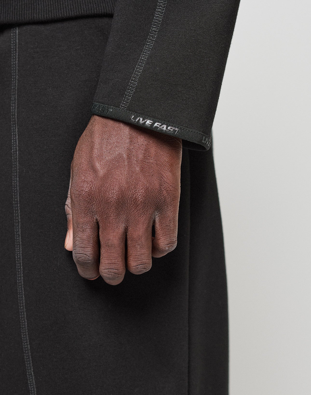 A close-up of a hand resting on black pants, wearing the LIVE FAST DIE YOUNG LFDY Active Fleece Hooded Zip in an Oversize Fit with LIVE FAST text on the sleeve cuff.