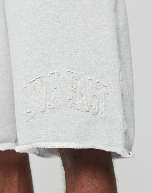 A close-up of the Ripped College Shorts from LIVE FAST DIE YOUNG, featuring LIVE FAST embroidered in white on the left leg, a raw unfinished hem, and a relaxed oversized fit for effortless style.