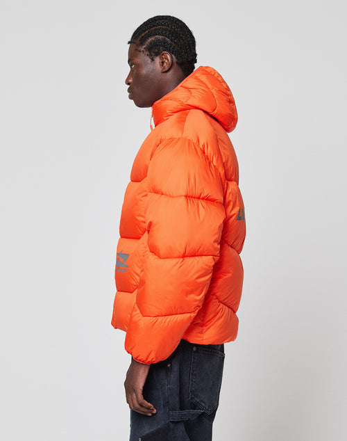 A person in profile wears the Live Fast Puffer Jacket by LIVE FAST DIE YOUNG—an orange, water-repellent hooded jacket—paired with dark jeans, standing against a plain, light gray background.