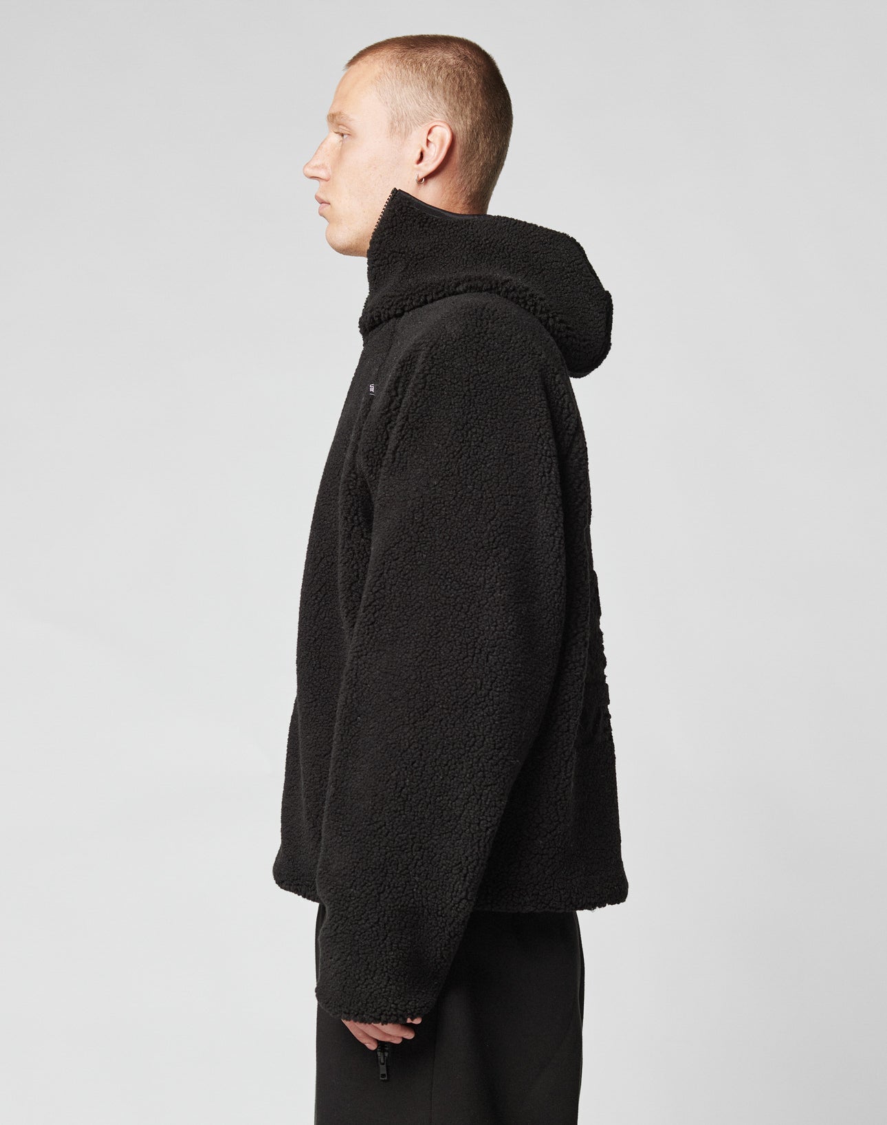 A person with short hair stands in profile, wearing the LIVE FAST DIE YOUNG Live Fast Teddy Jacket with a hood and black pants against a plain light gray background.