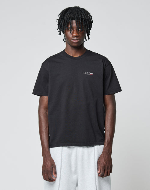 A person with short dreadlocks wears the LIVE FAST DIE YOUNG Live Fast Paris Tee, a black 100% Baumwolle T-shirt with small white text on the left chest, and light gray pants, standing against a plain light gray background.