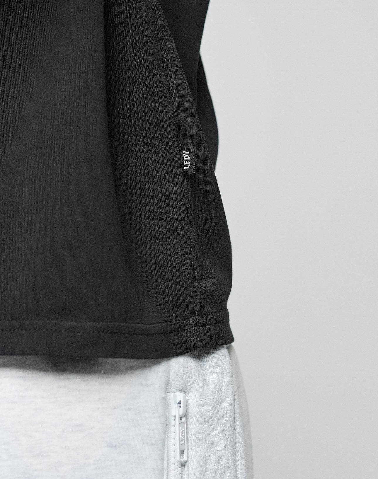 Close-up of a person wearing the LIVE FAST DIE YOUNG Live Fast Paris Tee in black with a small REP tag, made from 100% Baumwolle, paired with light gray pants featuring a white zipper pocket. Plain light background.
