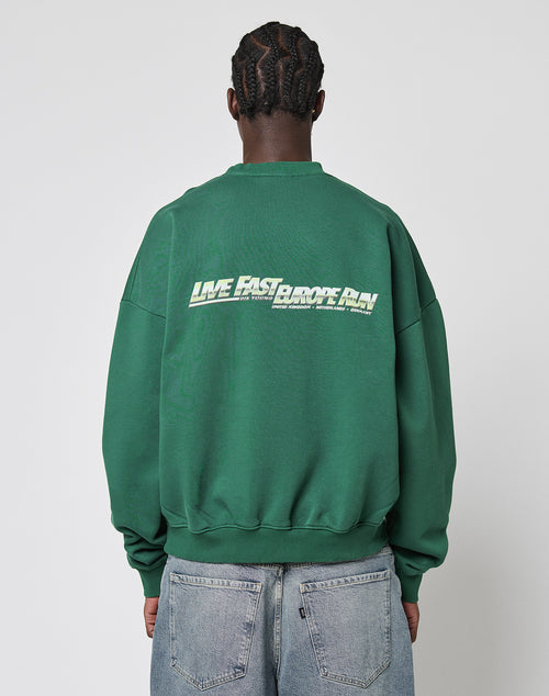 A person with braided hair, facing away, wears the LIVE FAST DIE YOUNG LFDY Europe Run Sweater in green with bold white LIVE FAST RACE AM text on the back, paired with light blue jeans.