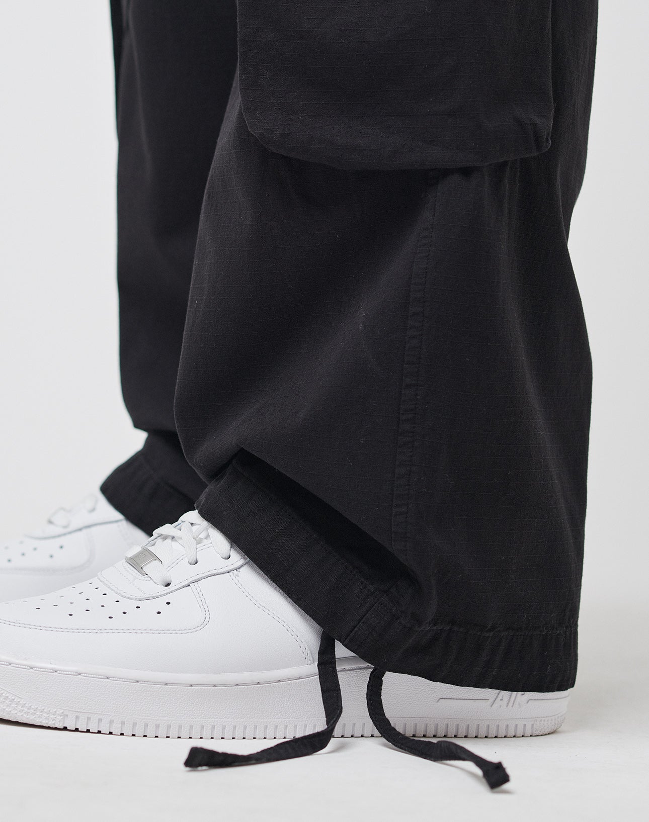 A close-up shows a person wearing LIVE FAST DIE YOUNG Wide Summer Cargo pants in black, featuring a loose fit, Cargotaschen, and drawstring hems draped over white Nike Air Force 1 sneakers against a plain white background.