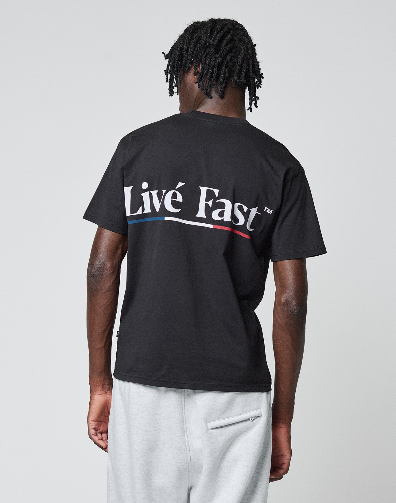A person with short, twisted hair faces away, wearing a black Live Fast Paris Tee by LIVE FAST DIE YOUNG—100% Baumwolle, front and back print “Live Fast” in large white letters—and light gray sweatpants, against a plain gray background.