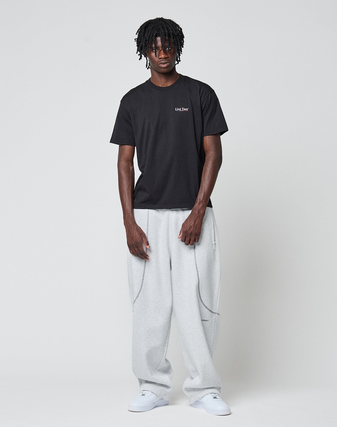 A man with short dreadlocks wears the LIVE FAST DIE YOUNG Live Fast Paris Tee with front and back print, light gray baggy sweatpants, and white sneakers, standing against a plain light gray background.