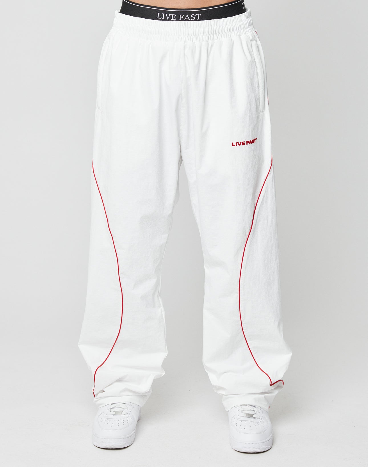 The LFDY Wavy Trackpants by LIVE FAST DIE YOUNG feature thin red piping, siliconeprint LIVE FAST text on the upper left thigh, zippered pockets, a black waistband with matching text, and pair perfectly with white sneakers.