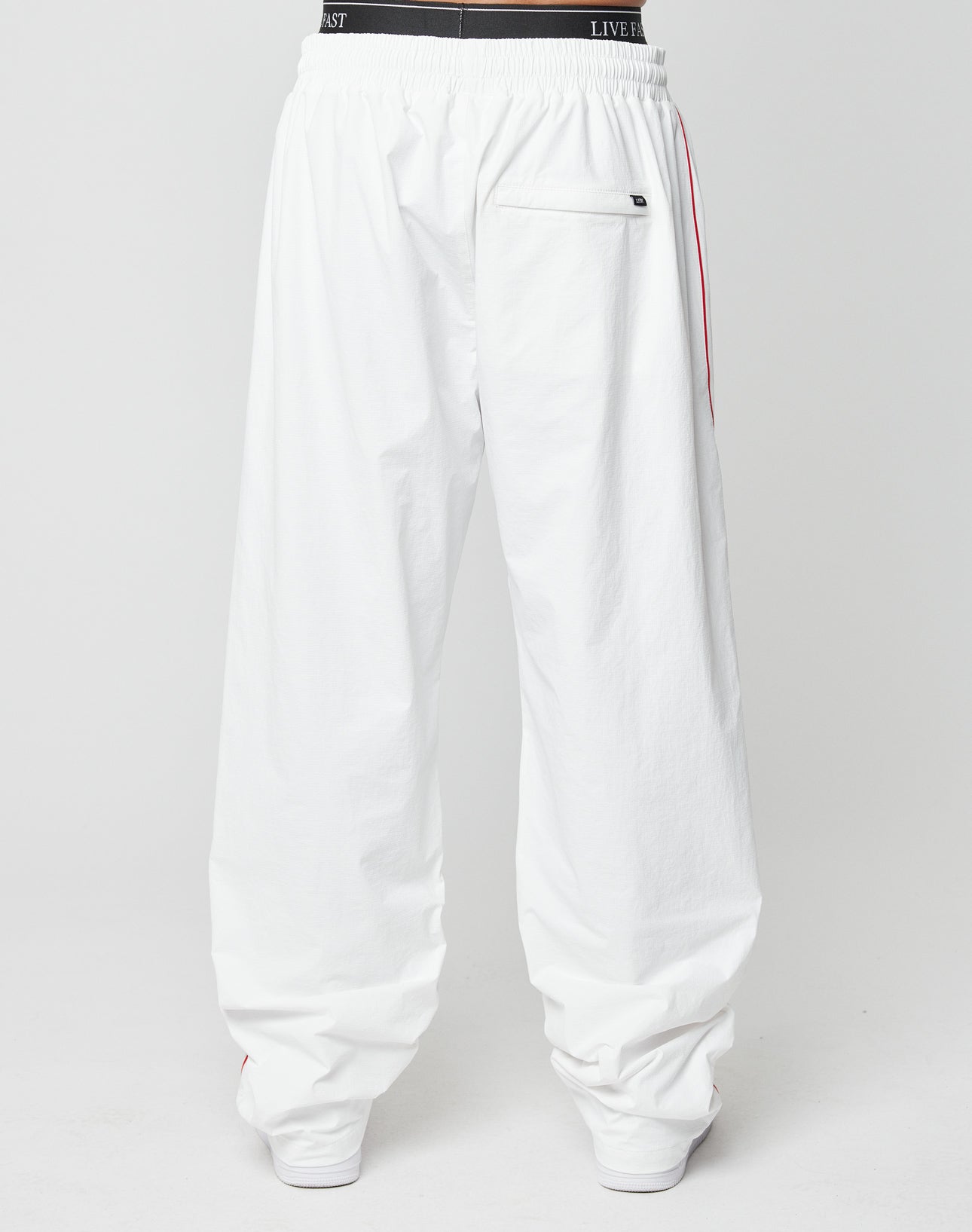 A person wearing LIVE FAST DIE YOUNG LFDY Wavy Trackpants—wide fit, loose white pants with an elastic waistband, visible back pocket with logo tag, and subtle red side stripes—stands against a plain white background.