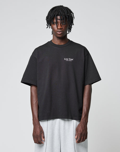 A person with short dreadlocks wears an oversize-fit black LIVE FAST DIE YOUNG LFDY Paris Tee made from 100% Baumwolle with “Live Fast” text on the chest, paired with light gray shorts against a plain light gray background.