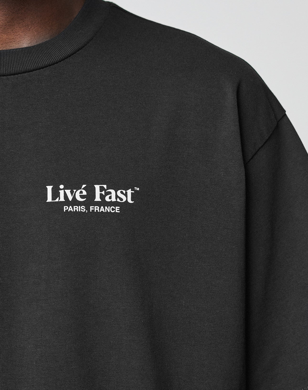 Close-up of a person wearing the LFDY Paris Tee by LIVE FAST DIE YOUNG. The black oversize fit features Livé Fast™ PARIS, FRANCE in white on the left chest. Made from 100% Baumwolle, the image highlights the neck and upper chest area.