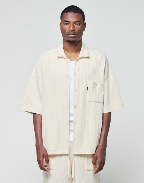 A man stands facing forward wearing the LIVE FAST DIE YOUNG Basic Knit Shirt in cream, an oversize, short-sleeve button-up with Hemdkragen and front pocket, paired with matching pants against a plain light background.