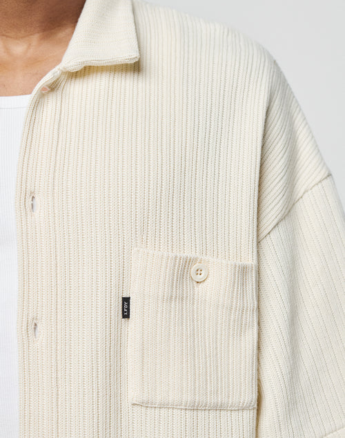A close-up shows a person wearing the LIVE FAST DIE YOUNG Basic Knit Shirt in cream with Hemdkragen, ribbed texture, front pocket, white inner shirt, and relaxed Oversize Fit. Only the chest and part of the neck are visible.