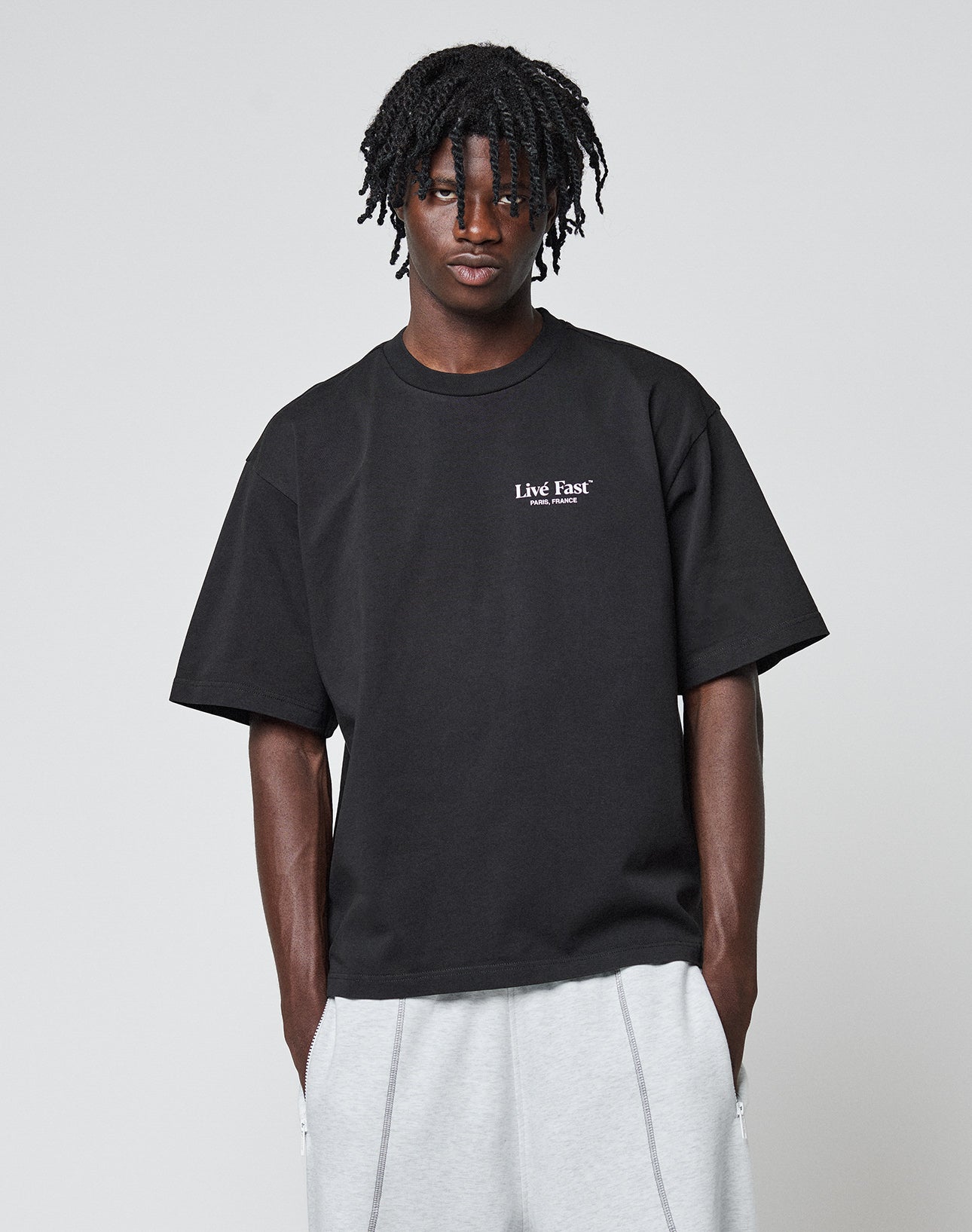 A person with medium-length, twisted hair wears an oversize fit black LFDY Paris Tee by LIVE FAST DIE YOUNG and light gray sweatpants, standing against a plain light gray background.