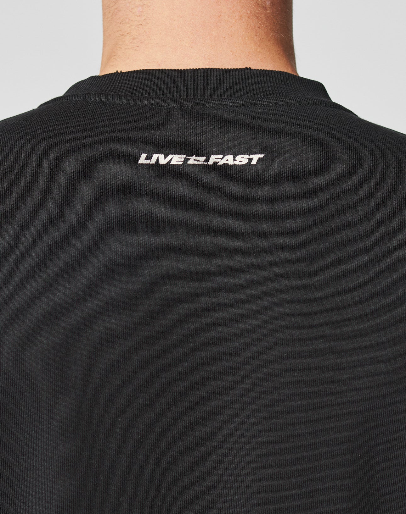 The back of the LFDY Mystic Sweater by LIVE FAST DIE YOUNG, featuring black ribbed cotton and “LIVE FAST” in white near the neckline. The clean topstitching is visible above the wearer’s neck and partial head.