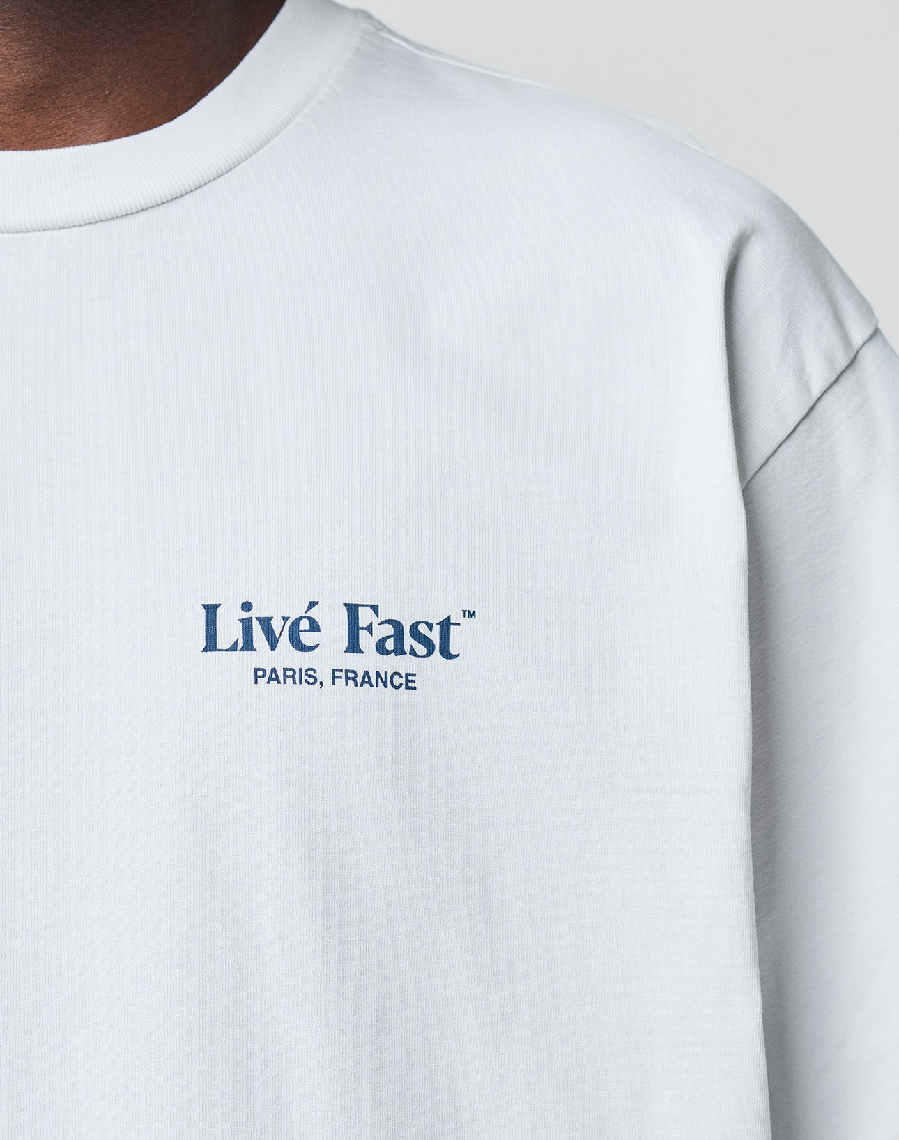 A close-up of the LIVE FAST DIE YOUNG LFDY Paris Tee in white Baumwolle shows Livé Fast™ PARIS, FRANCE in blue on the left chest. Only part of the neck and shoulder are visible, emphasizing its Oversize Fit.