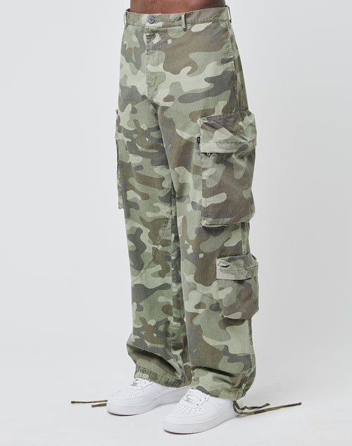 A person wearing LIVE FAST DIE YOUNG Army Cargo wide fit camouflage cargo pants with multiple pockets and white sneakers, standing against a plain white background.
