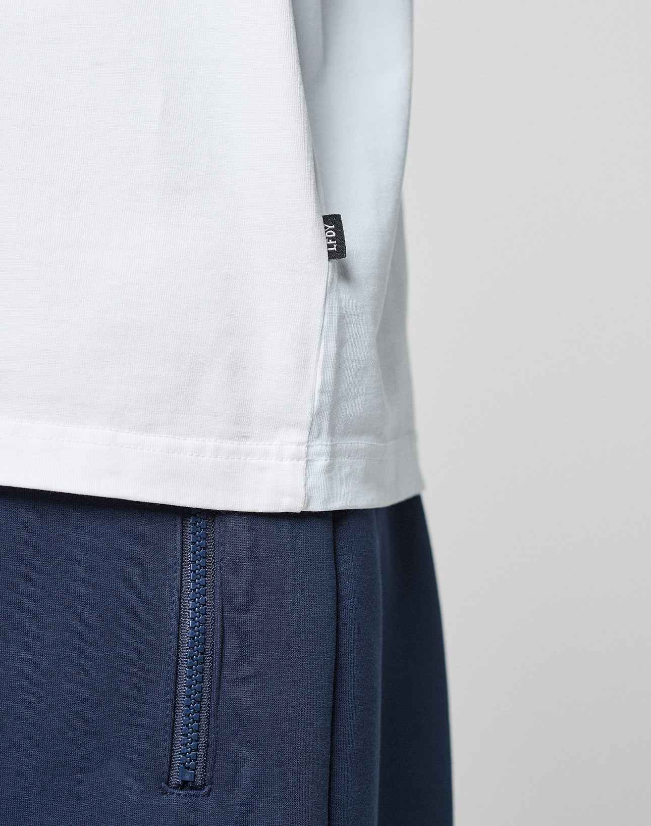 Close-up of a person wearing the LFDY Paris Tee by LIVE FAST DIE YOUNG in white, featuring a small black tag on the side seam, styled with blue pants with a visible zipper pocket. The background is plain and light-colored.