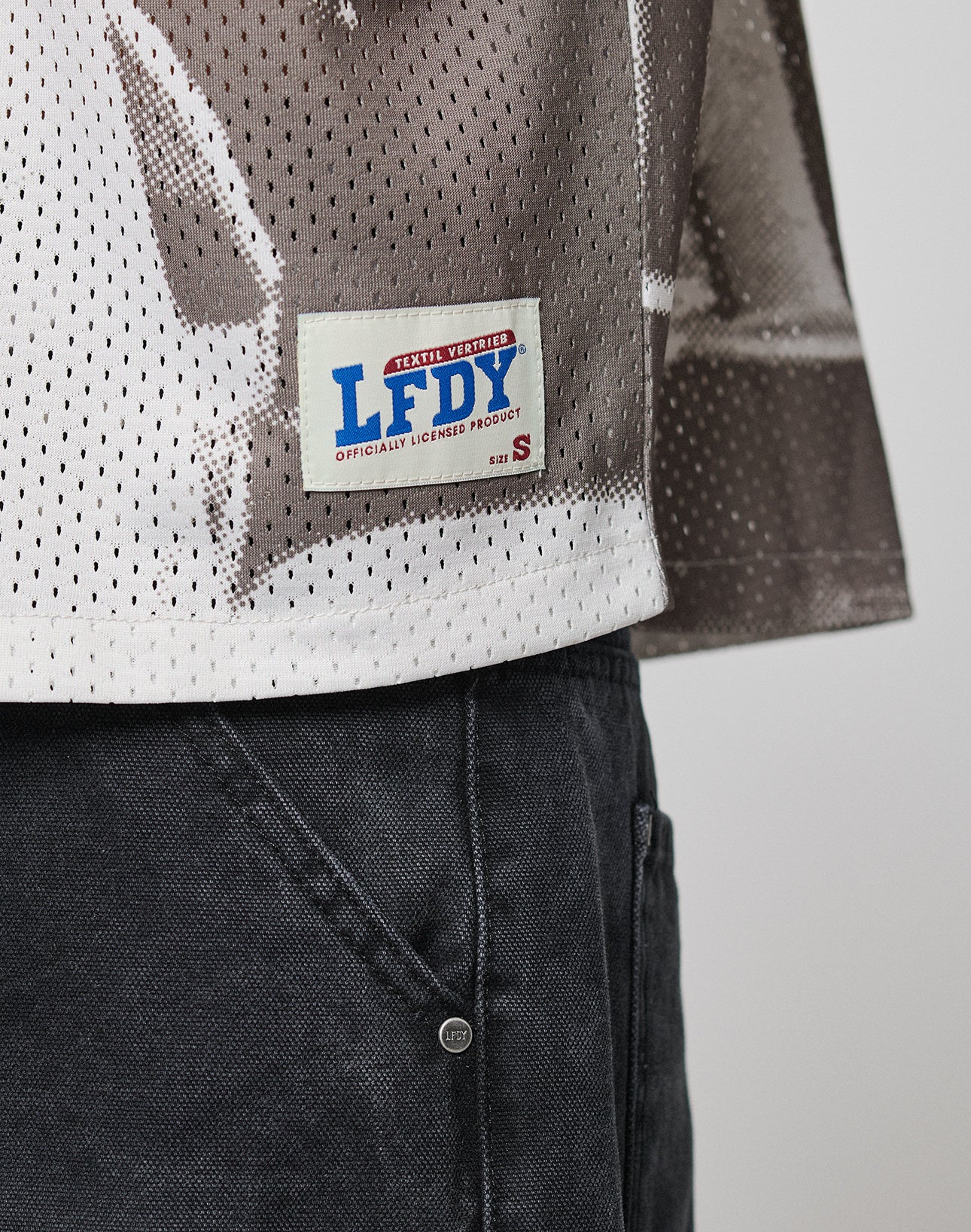 A close-up shows the lower part of the oversize LFDY Courtroom Jersey by LIVE FAST DIE YOUNG, featuring perforated patterned fabric and a sewn patch labeled LFDY OFFICIALLY LICENSED PRODUCT, paired with dark gray pants.