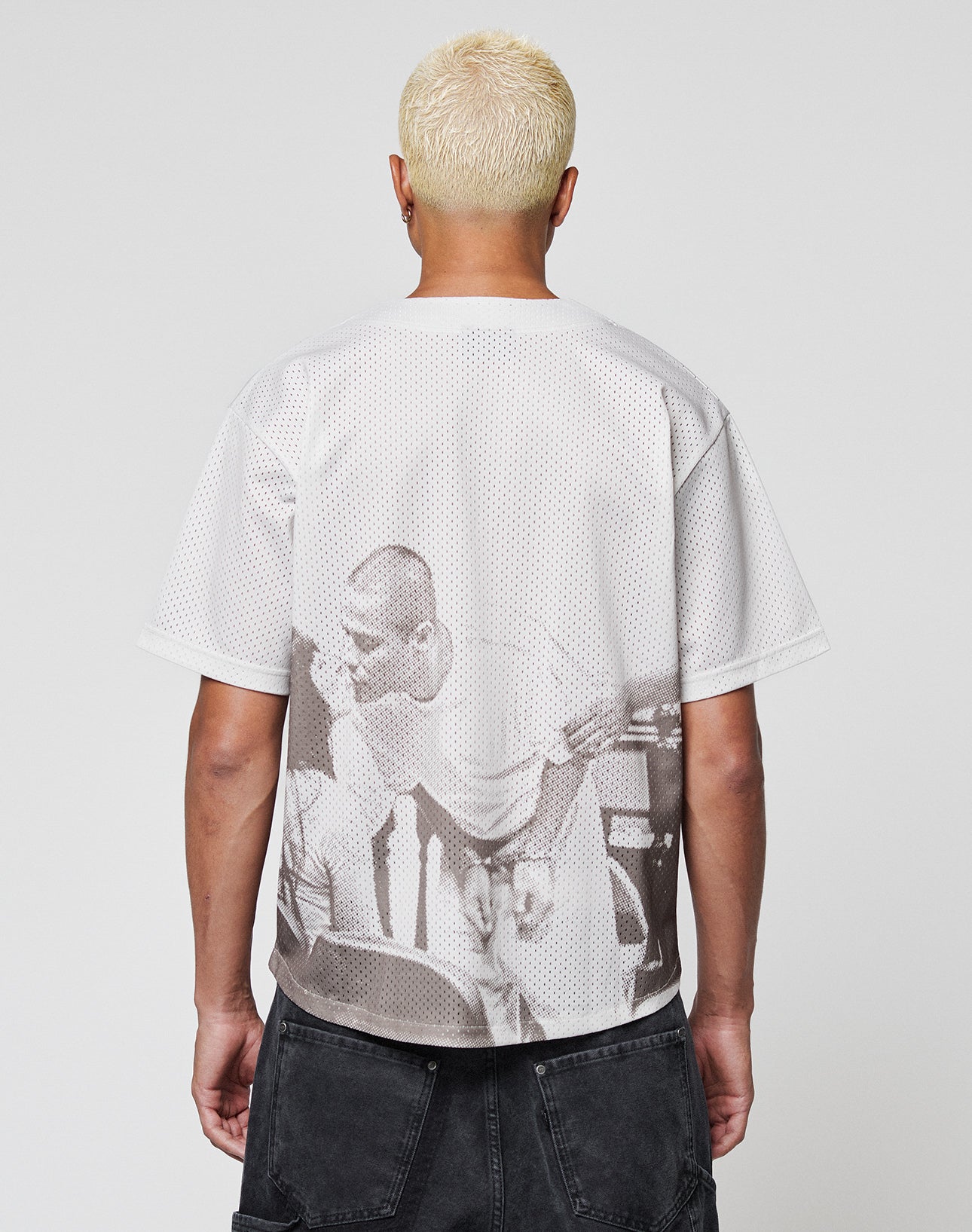 A person with short, bleached blond hair faces away, wearing the oversize LIVE FAST DIE YOUNG LFDY Courtroom Jersey featuring a sepia image on the back, paired with dark shorts against a plain, light background.