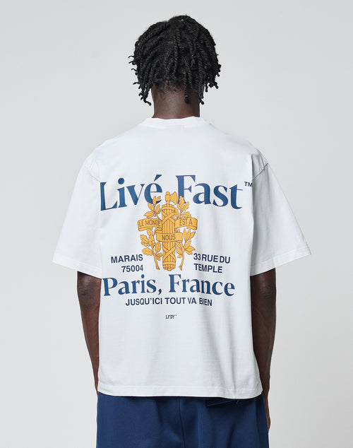 A person wearing the LFDY Paris Tee by LIVE FAST DIE YOUNG, an oversized white cotton T-shirt with blue and yellow front and back graphics reading “Live Fast™ Paris, France” and French text, stands against a plain light background.