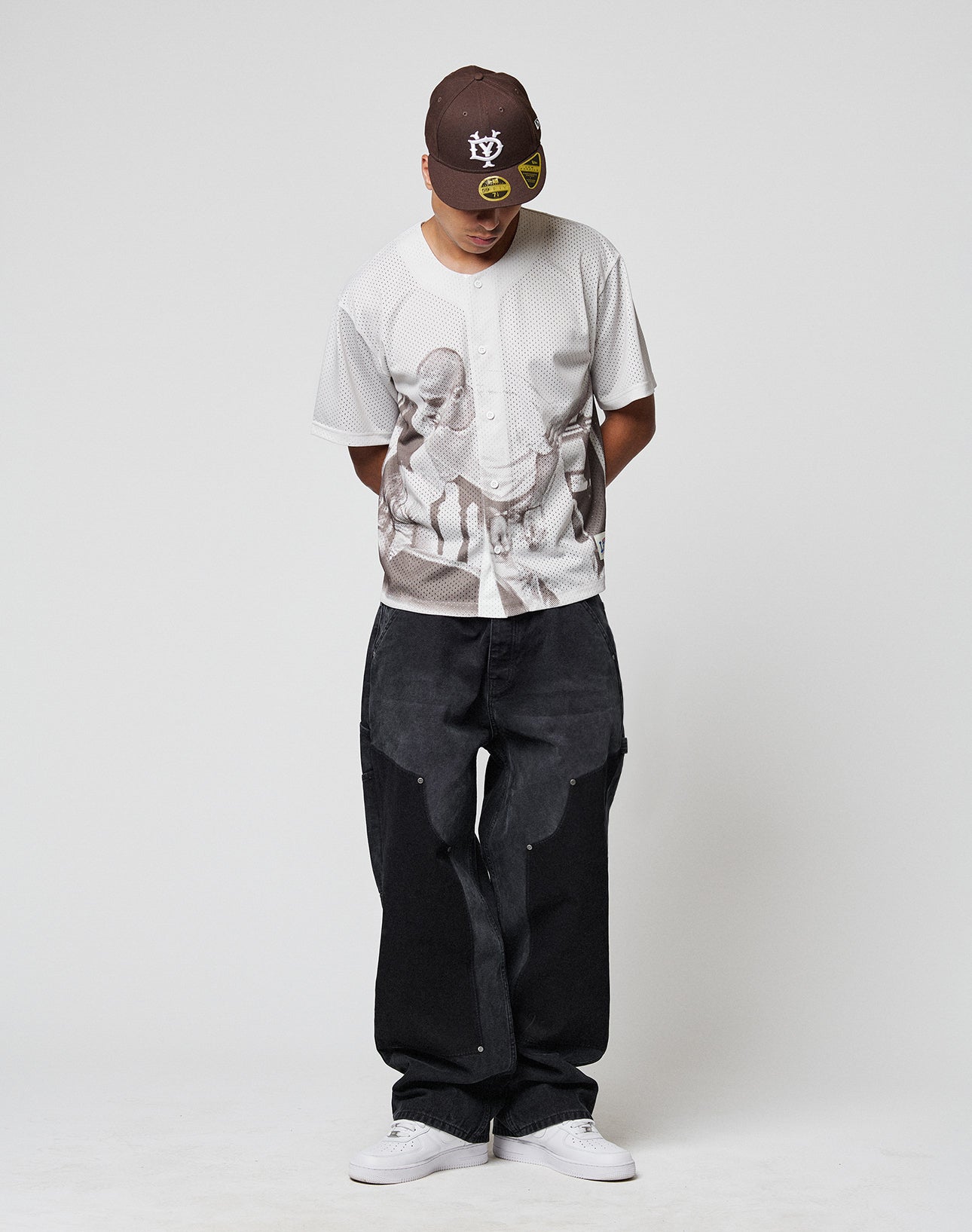 A person wearing a brown baseball cap, loose black jeans, white sneakers, and the LIVE FAST DIE YOUNG LFDY Courtroom Jersey stands against a plain white background, looking down with hands behind their back.