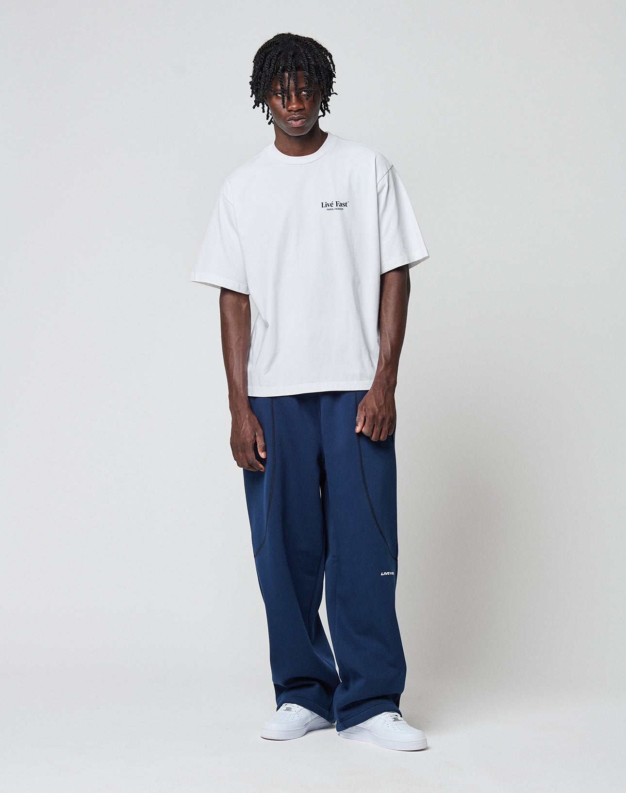 A person stands against a plain background wearing a loose white T-shirt with a small logo on the left chest, LFDY Active Fleece Jogger by LIVE FAST DIE YOUNG in navy blue, and white sneakers.