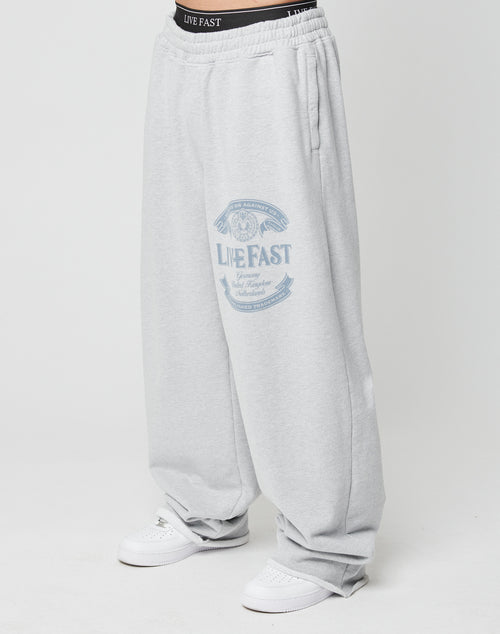 A person wears Loyal Wide Jogger sweatpants by LIVE FAST DIE YOUNG in light gray, featuring a wide leg, elastic waistband, and graphic on the upper thigh. Black underwear is visible above the pants, paired with white sneakers.