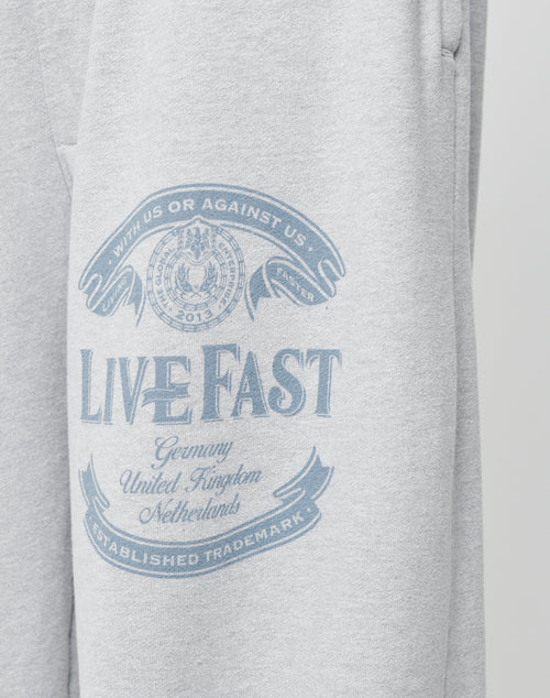 The Loyal Wide Jogger by LIVE FAST DIE YOUNG features light gray fabric with a blue LIVE FAST print, banners for Germany, UK, Netherlands, and Established Trademark. It offers an oversize fit and elastic waistband for extra comfort.