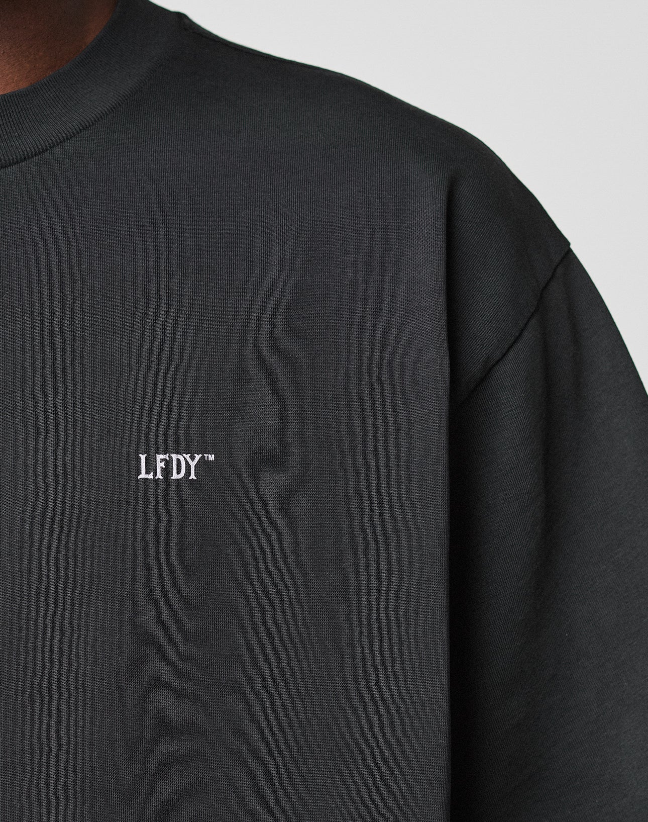 Close-up of a person wearing the LIVE FAST DIE YOUNG Basic Trademark Tee. The black crewneck features white embroidered LFDY™ on the chest. Only part of the shoulder, neck, and upper chest is visible. Made from 100% Baumwolle.