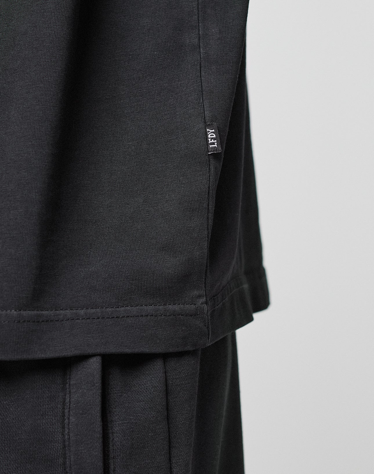 Close-up of a person wearing the LIVE FAST DIE YOUNG Basic Trademark Tee in black with an oversize fit, paired with black pants. A small black ACGT tag is visible at the hem. The background is plain and light-colored.