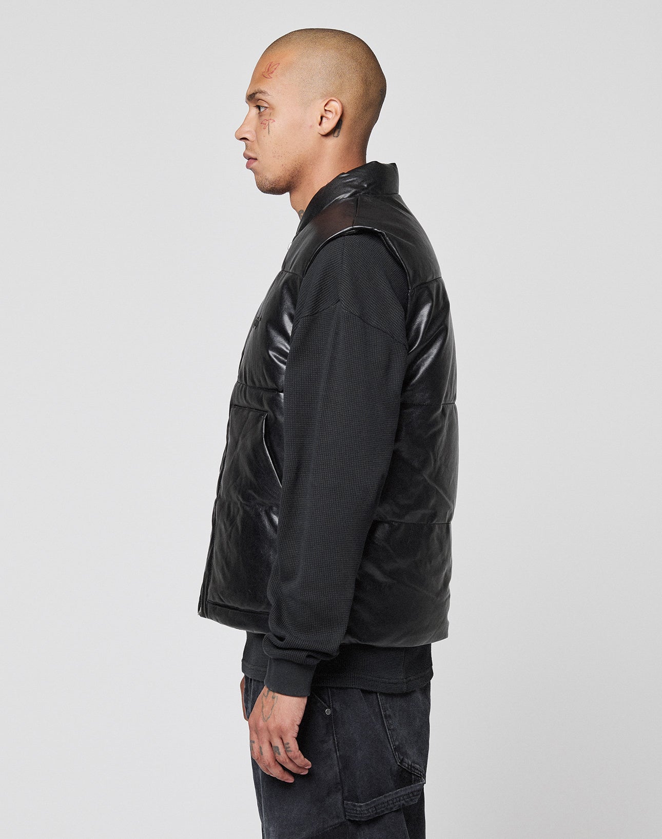 A man with a shaved head stands in profile against a plain background, wearing a black long-sleeve shirt, dark pants, and a regular fit Padded Vest with padded stand-up collar by LIVE FAST DIE YOUNG.