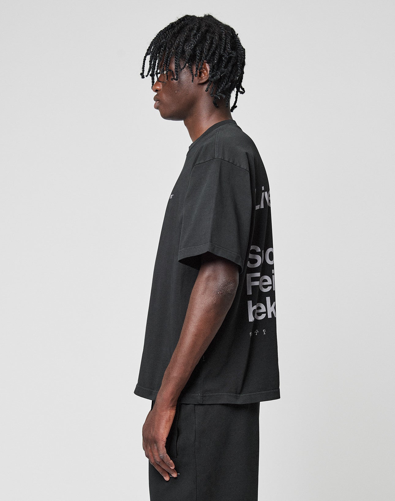 A person with short, twisted hair stands in profile against a plain light background, wearing the LIVE FAST DIE YOUNG Basic Trademark Tee—an oversized fit black t-shirt made from 100% cotton with bold front and back prints—and matching black pants.