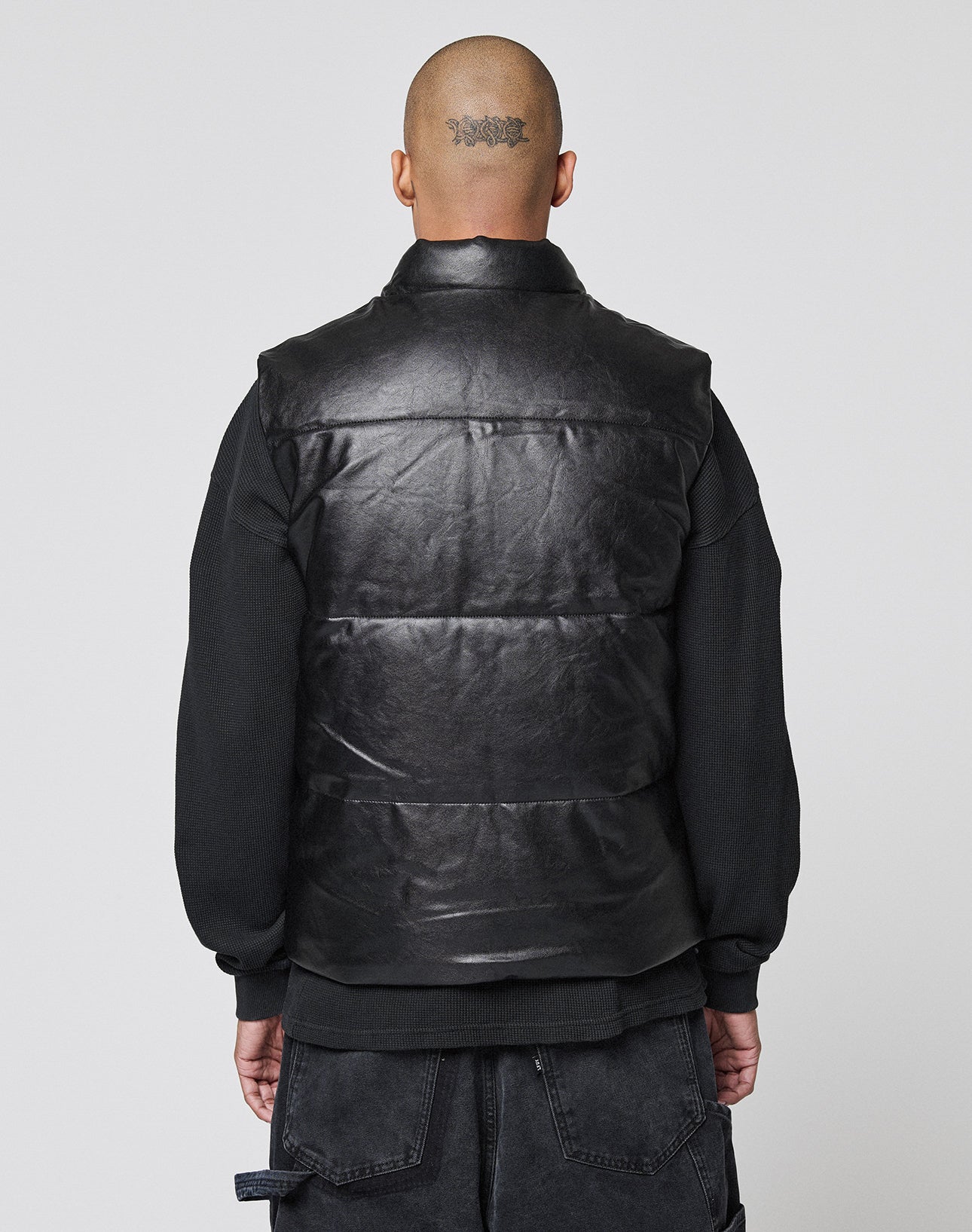 A person with a shaved head and a head tattoo is seen from behind, wearing the LIVE FAST DIE YOUNG Padded Vest in black over a black sweatshirt, standing against a plain light background.